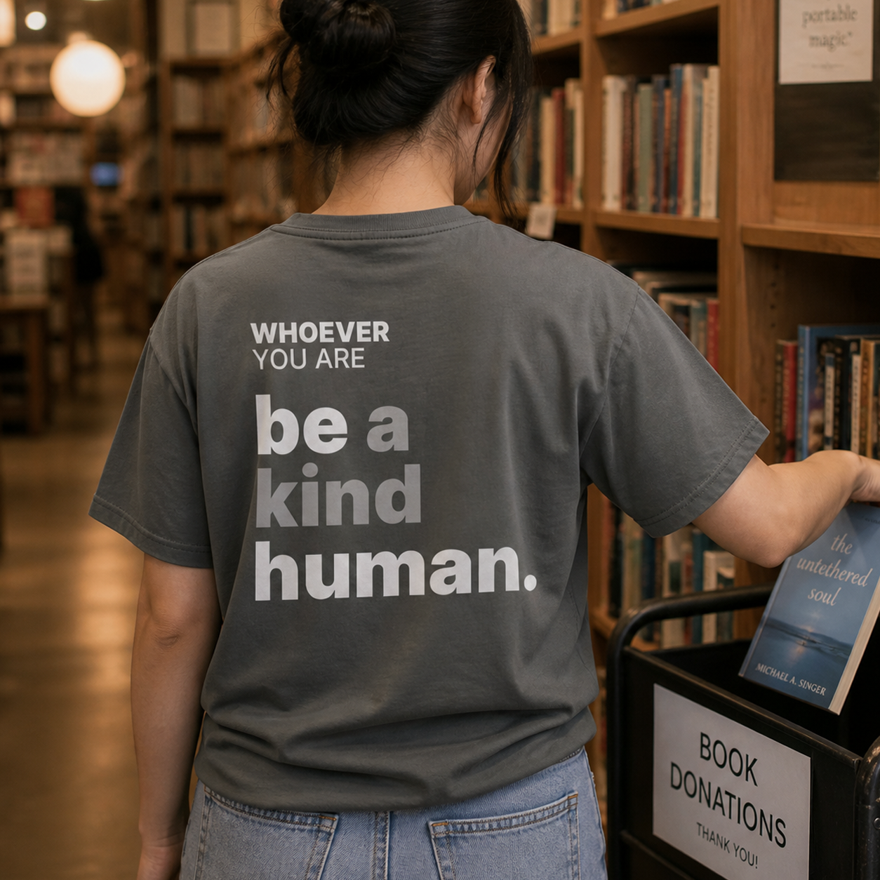 Person wearing a gray Be a Kind Human kindness t-shirt in a bookstore setting